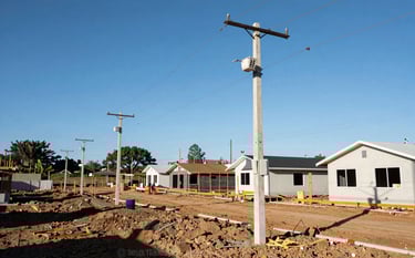 Wide shot of a South American residential allotment under construction, showing newly installed utility poles and clean electrical infrastructure against a bright blue sky. Professional and efficient site organization.