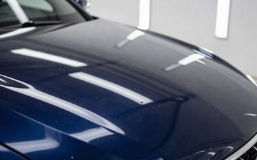 Macro photography of a deep navy blue car hood in a North American studio. The paint reflects a perfect, mirror-like image of the overhead lights after a professional ceramic coating. The gloss is deep and liquid-like, showcasing ultimate precision.