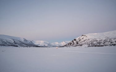 A wide, calming landscape of a snow-covered valley in the Arctic Circle under a soft blue twilight. A single small campfire glow is visible in the distance. The composition is minimalist and vast, emphasizing the quiet power of winter. Soft contrasts with colors #5E4B3E and #F4F0E4.