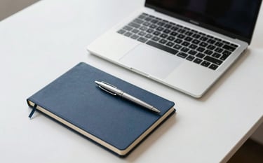 A minimalist, high-end office desk setup featuring a slim laptop on a crisp white surface. A deep navy blue notebook and professional silver pen sit nearby. The lighting is bright and airy, suggesting a modern and innovative workspace.