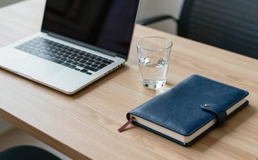 A clean and organized office desk in a European Portuguese setting. A sleek laptop, a leather-bound notebook, and a glass of water are positioned on a minimalist wooden surface. Soft natural morning light illuminates the scene, featuring subtle accents of light blue and dark blue. Professional and efficient atmosphere.