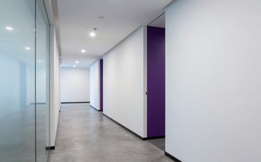 A crisp, professional wide-angle shot of a minimalist North American office hallway. The walls are clean white, with glass dividers reflecting soft blue and deep purple accent lighting.