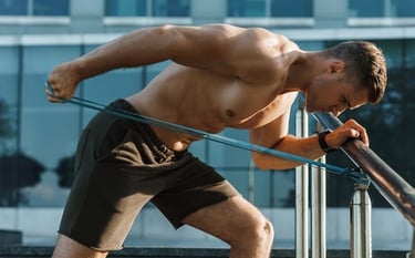 Shirtless muscular man performing tricep kickbacks with a resistance band outdoors for strength training.