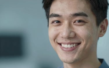 A close-up portrait of a person showing a confident, radiant, and healthy white smile. The background is softly blurred with tones of muted blue (#4A6572), conveying the high standard of aesthetic dentistry results.