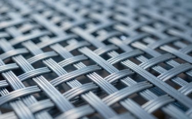A professional industrial shot of heavy-duty manganese steel woven screens. The focus is on the intricate and robust weaving pattern of the wire. The composition is dynamic and diagonal, showcasing the strength of the material. The lighting reflects off the metallic surfaces with cool tones matching #A3B5B5 and #2C3A3B.