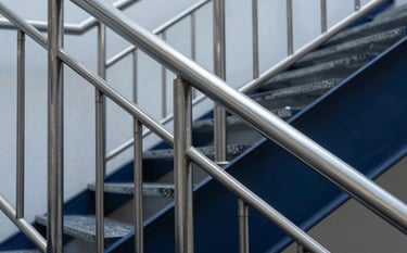 Close-up architectural shot of a precision-engineered stainless steel railing and staircase in a contemporary South American / Brazilian commercial building. The steel blue and dark navy reflections of the metal are sharp. The lighting is studio-quality, emphasizing the brushed texture and professional finish of the inox projects.