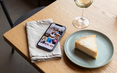 A flat lay photography shot on a light wood Scandinavian-style table in a North American cafe. A smartphone showing a vibrant social media feed sits next to a linen napkin, a matte green ceramic plate with artisanal cheese, and a glass of wine. Sophisticated, bright natural lighting, professional and clean composition.
