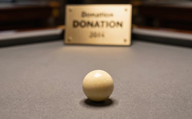 A sophisticated close-up of a pool table with a donation plaque visible in the blurred background. The lighting is warm and soft, emphasizing the professional felt texture and muted gold billiard accessories.