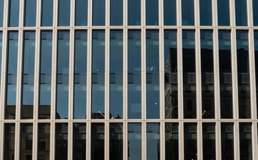 Minimalist architectural photography of a modern glass office building facade in a North American city. Strong geometric lines, deep blue sky reflections, and a sense of absolute precision. Soft natural lighting, off-white and deep navy tones.