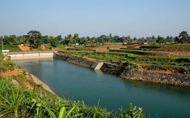 A wide-angle landscape photograph of a sustainable watershed project in a South Asian / Indian rural region. Stone check dams and terraced hillsides are visible under a clear sky. The colors include deep teal water and vibrant green vegetation.