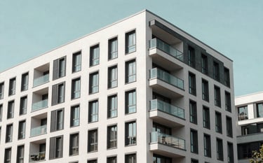Professional architectural photography of a modern apartment complex in Berlin. The building features clean lines, large glass balconies, and a facade in soft off-white and deep charcoal accents. The sky is a clear pale seafoam color, indicating a bright day.