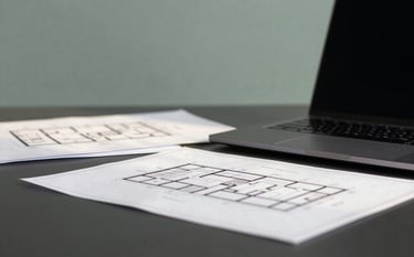 A close-up of a high-end office desk in Berlin. A laptop and architectural plans are neatly arranged on a deep charcoal surface. In the background, a muted sage green wall provides a professional backdrop. Soft, morning sunlight pours in.