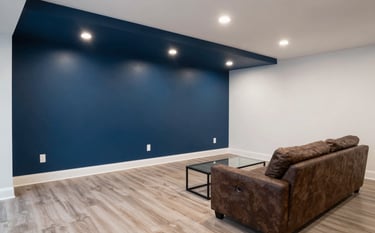 A newly finished basement interior in a North American / Canadian (with South Asian cultural influence) home. The room is styled as a cozy media lounge with a steel blue accent wall and professional recessed ceiling lights. High-quality flooring and clean white walls emphasize the renovation quality.