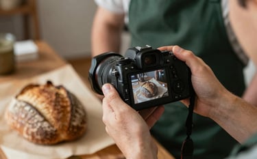 A behind-the-scenes close-up of a digital marketer's hands holding a professional camera, capturing a rustic sourdough loaf on #FDF5E6 parchment paper. The photographer wears a #364536 forest green apron. Sophisticated, warm lighting.