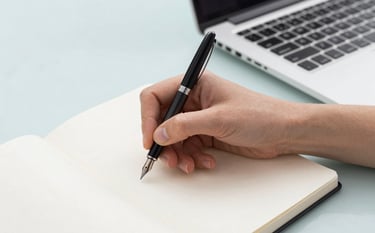 Close-up of a person's hand using a fountain pen on a white notebook next to a high-end laptop on a Pale Azure desk. The aesthetic is clean and minimalist, representing strategic planning and focus. Lighting is bright and even.