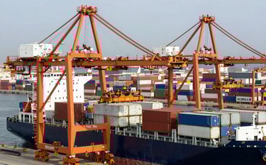 A high-angle professional photograph of a bustling European / Spanish cargo port. Large vibrant orange shipping cranes are loading white containers onto a massive cargo ship. The scene is bathed in clear daylight, emphasizing efficiency and modern logistics.