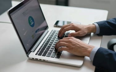 A close-up of a person's hands in a modern North American office, typing on a clean silver laptop. Beside the laptop is a smartphone showing a secure notification badge. The desk is organized and professional, with cool steel blue and off-white lighting.