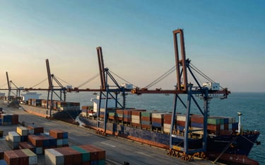 A high-angle wide shot of a bustling modern Middle Eastern / Gulf shipping port at sunrise. Huge container ships are docked, with towering industrial cranes silhouetted against a soft teal and deep ocean blue sky. The scene conveys robust commercial activity and professional logistics.