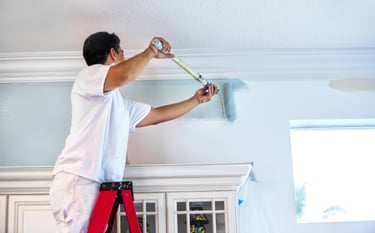 Professional house painter on a ladder using a roller to apply light blue paint to an interior wall.