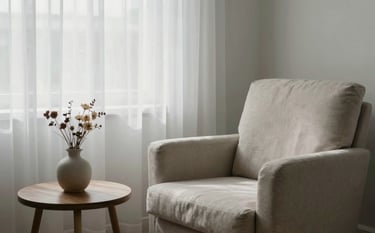 Photography of a peaceful therapy room corner. A soft-textured armchair sits by a window with misty white curtains. On a side table, there is a small ceramic vase with dried flowers. The lighting is soft and natural, featuring tones of misty white and soft sage.