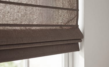 Close-up photography of custom Roman shades in a muted brown fabric, installed in a bright North American breakfast nook. The lighting is crisp and natural, showcasing the precise fit and clean lines of the blinds against a soft white wall.