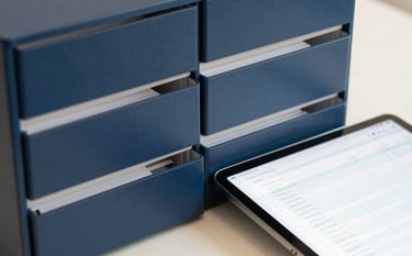 A close-up photograph of a organized physical filing system and a tablet showing a neat digital spreadsheet. The composition is clean and focused, using a color palette of midnight blue and cool off-white to emphasize efficiency and meticulous document organization.