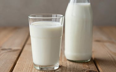 A close-up photograph of a fresh, cold glass of milk next to a glass bottle on a rustic wooden table. The scene is bright and highlights purity and health. The background is softly blurred to emphasize the product. Colors: soft cream and warm taupe.