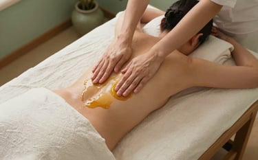 A top-down photographic view of a serene spa setting. A person lies on a wooden table covered in soft off-white linens. Warm, golden herbal oil is being gently massaged into their back by hands in a rhythmic motion. The room is decorated with sage green accents and soft moss colored ceramics. Professional, tranquil lighting.