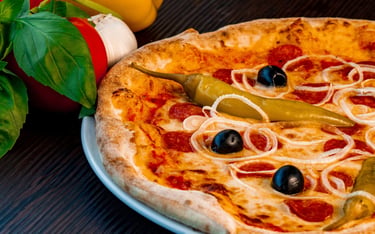 Steinofen-Pizza mit Salami, Oliven, Zwiebelringen und Peperoni neben frischem Basilikum.