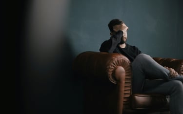A stressed man sitting on a brown leather armchair holding his head in frustration.