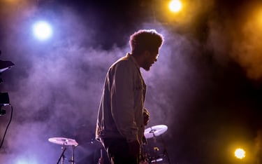 A male musician performs on a smoky stage under purple and yellow concert lights.