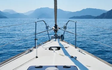 A white sailing yacht bow navigating blue ocean waters toward coastal mountains on a sunny day.