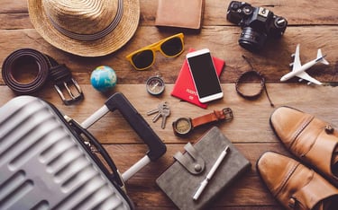 Travel essentials flat lay on a wooden table including a suitcase, passport, camera, and straw hat.