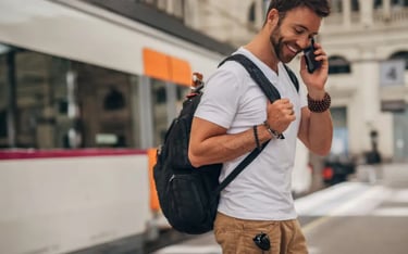 A smiling man with a backpack talking on a smartphone while waiting for a train.
