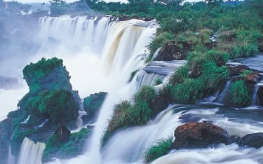 Panoramic view of Iguazu Falls with powerful white water cascading over green cliffs in a tropical rainforest.