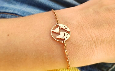 A gold world map charm bracelet worn on a wrist against a blue denim and yellow shirt background.