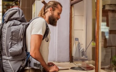 Male backpacker with a beard buying tickets at a train station counter during a solo travel trip.