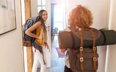 Two female backpackers with travel gear entering a bright hostel room for their budget adventure trip.
