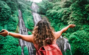 A female traveler with a red backpack overlooks a scenic tropical waterfall in a lush green jungle.