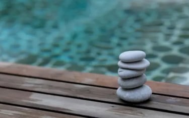 Balanced stack of smooth grey zen stones on a wooden deck by a tranquil blue swimming pool.