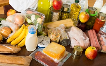 Variety of groceries including fresh fruit, vegetables, meat, cheese, and canned goods on a wooden table.