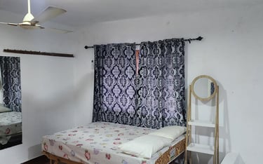 A small bedroom featuring a bed with patterned sheets, blue curtains, a ceiling fan, and a mirror.