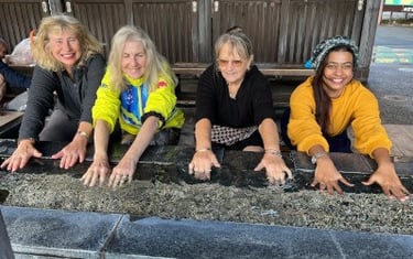 Four women seated, leaning forward with their hands in a long water tank with running spring water.