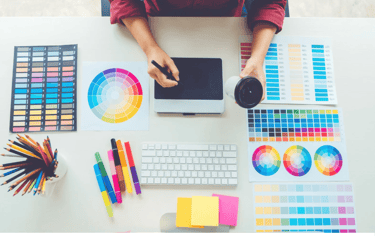 A graphic designer works at a desk with a digital drawing tablet, color palettes, and art supplies.