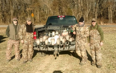 Four hunters in camouflage standing behind a truck tailgate displaying a harvest of Canada geese in a grassy field.