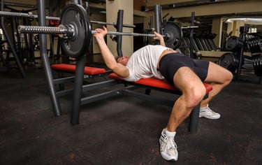 A muscular man performing a barbell bench press on a weight bench in a commercial gym setting.