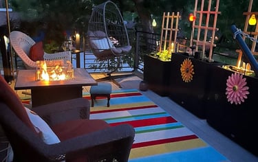 Cozy rooftop deck with gas fire pit table, colorful striped rug, and hanging egg chair at twilight.