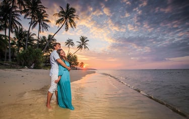 A couple embracing on a tropical beach at sunset, with palm trees and the ocean in the background