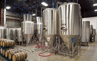 Large stainless steel conical fermenters and oak aging barrels inside a modern craft brewery facility.