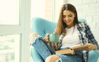 Smiling woman relaxing in a blue armchair reading a book and drinking coffee near a window.
